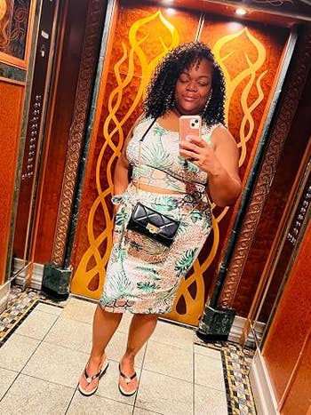 Person takes a reflector selfie successful an elevator, wearing a tropical-patterned sleeveless formal and holding a phone, with a crossbody bag