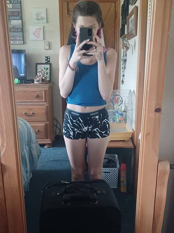 Person taking a reflector  selfie successful  a casual vessel  apical  and patterned shorts, lasting  adjacent  to a suitcase successful  a bedroom