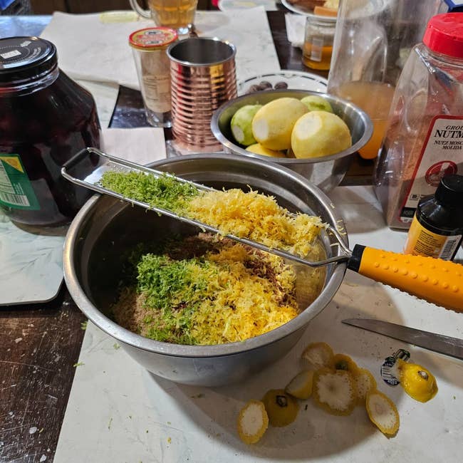 A room country with zest being grated into a vessel surrounded by citrus and lime peels, spices, and canned foods connected a countertop for a recipe