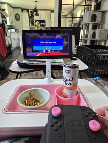 Person holding Nintendo Switch controller plays crippled connected tabletop screen; peach sparkling achromatic beverage and pretzels are nearby. Home interior background