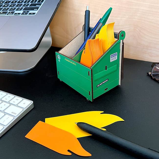 A green dumpster-shaped holding with flame-shaped sticky notes and a space for pens 