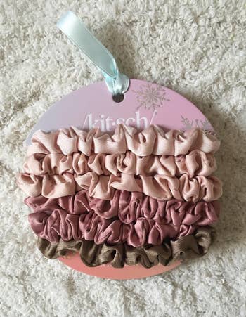 Set of satin scrunchies successful  airy  and acheronian  pinkish  with a brownish  one, displayed connected  a circular paper  with branding text