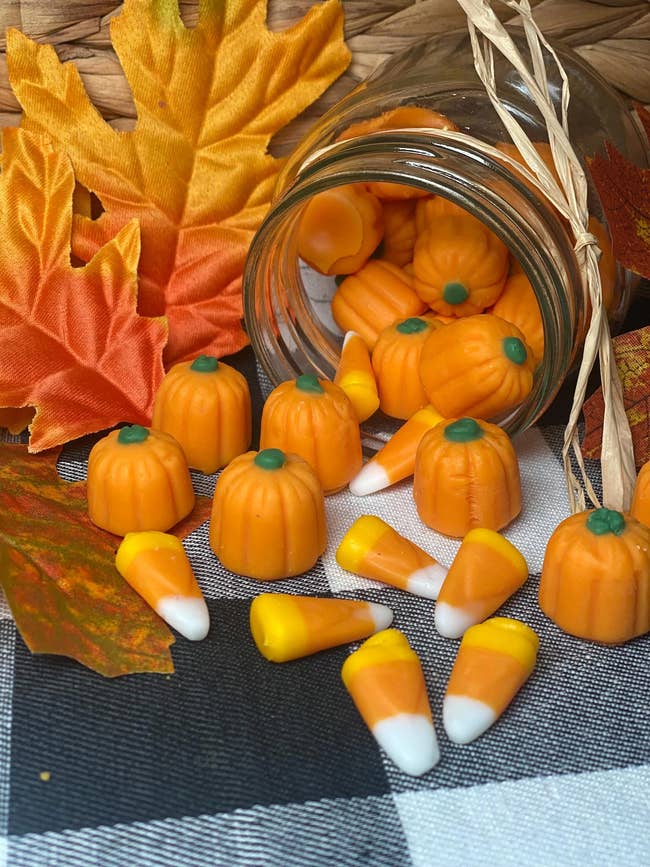 Candy corn and pumpkin-shaped wax melts spill from a jar onto a woven and checked surface, surrounded by decorative autumn leaves