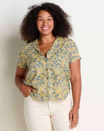 Woman smiling in a floral blouse and cream pants, hand on hip, for a shopping article
