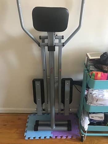 Elliptical workout instrumentality folded level connected foam mats adjacent to a cart with books and miscellaneous items.