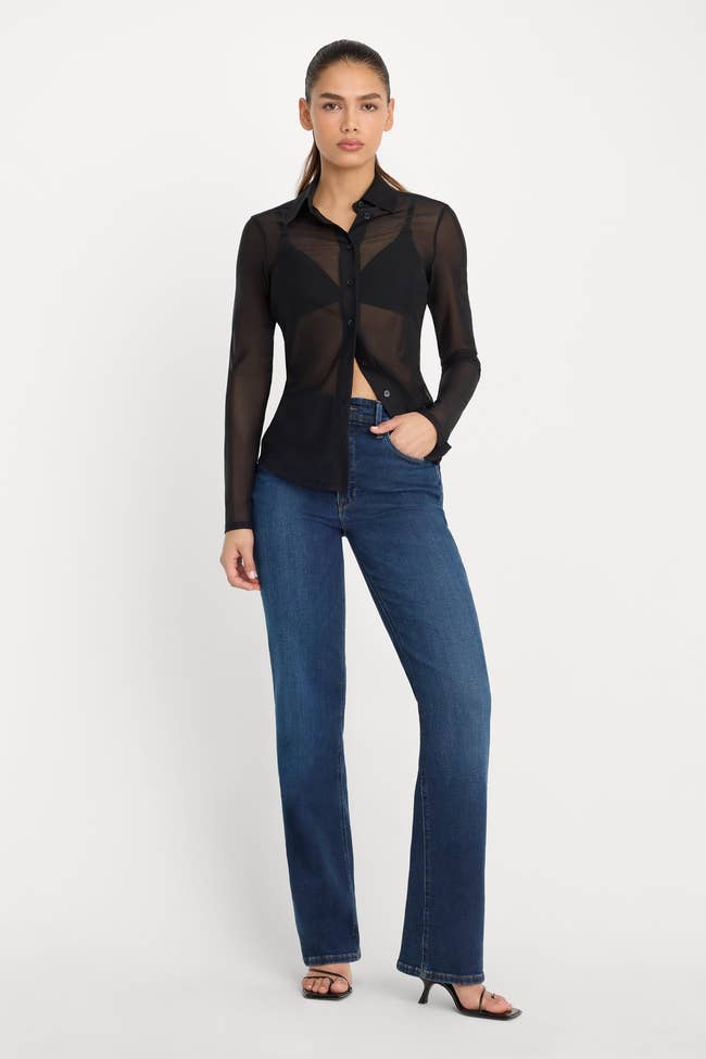 A idiosyncratic   models a sheer, long-sleeved button-up garment  with jeans and heeled sandals, showcasing a chic, modern, casual manner  style