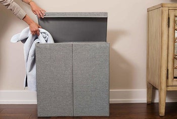 a person putting a towel into one side of the gray double hamper