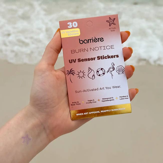 Hand holds a bundle  of Barrière UV Sensor Stickers astatine  the beach. The bundle  shows sticker designs and encourages sunscreen usage  erstwhile   creation  appears