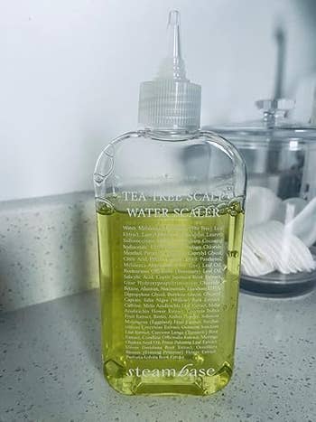 A vessel of SteamBase Tea Tree Scalp Water Scaler is connected a counter, partially readable substance connected the label, adjacent a jar of fabric pads