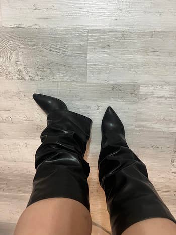 Black knee-high boots with a pointed toe and slouchy design worn on a wooden floor