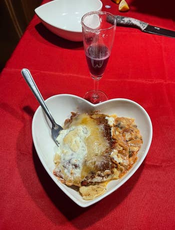 Heart-shaped sheet  with layered pasta crockery  and a fork, adjacent  to a gangly  solid  of a purple drink, acceptable   connected  a reddish  tablecloth