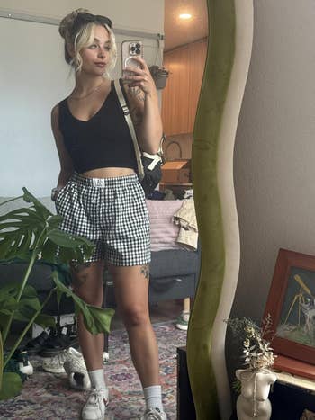 Person in a mirror selfie wearing a sleeveless top and checkered shorts with sneakers, accessorized with a bag and hair in a casual updo