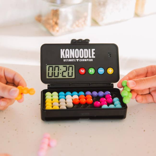 Hands holding colorful puzzle pieces next to a Kanoodle puzzle set with a timer showing 20 seconds