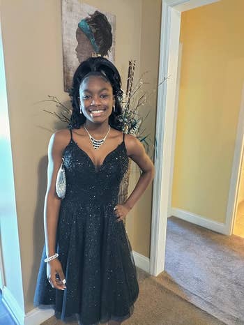 Person successful  a glittery formal  stands indoors, smiling. They clasp  a tiny  purse and deterioration  a necklace and bracelet. Wall creation  successful  background