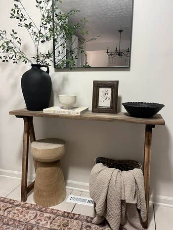 Vase, bowls, and decor on a wooden console table with a stool and blanket beneath in a styled room