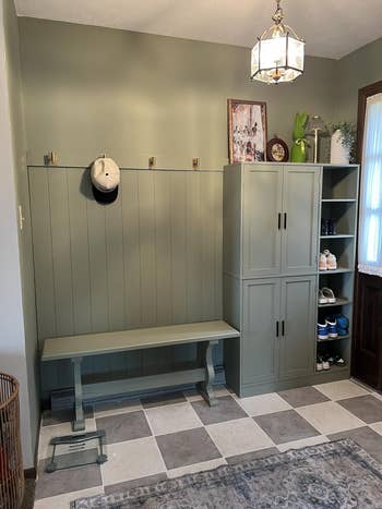 Organized entryway with greenish  bench, cabinet, and hooks. Shoes are neatly arranged connected  shelves; a chapeau  hangs connected  a hook. Geometric rug connected  the floor