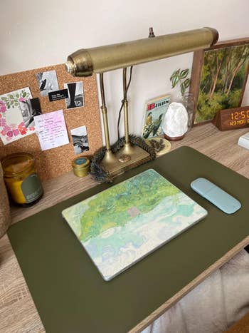 A corkboard table  setup with a golden  lamp, greenish  table  mat, laptop, wireless mouse, candle, plant, clock, and assorted notes and photos pinned to the board