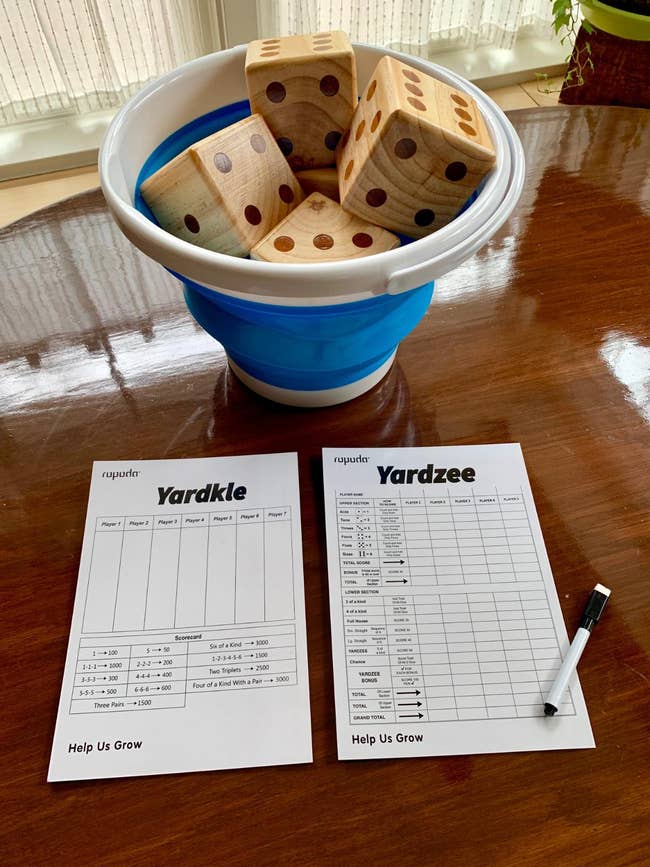 A set of Yardzee and Yardkle score sheets on a wooden table, with a collapsible bucket containing large wooden dice. A black pen is placed next to the sheets