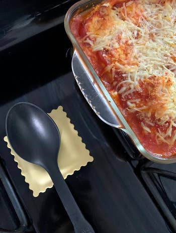 A casserole dish with lasagna next to a spatula on a stovetop resting on a silicone ravioli rest 