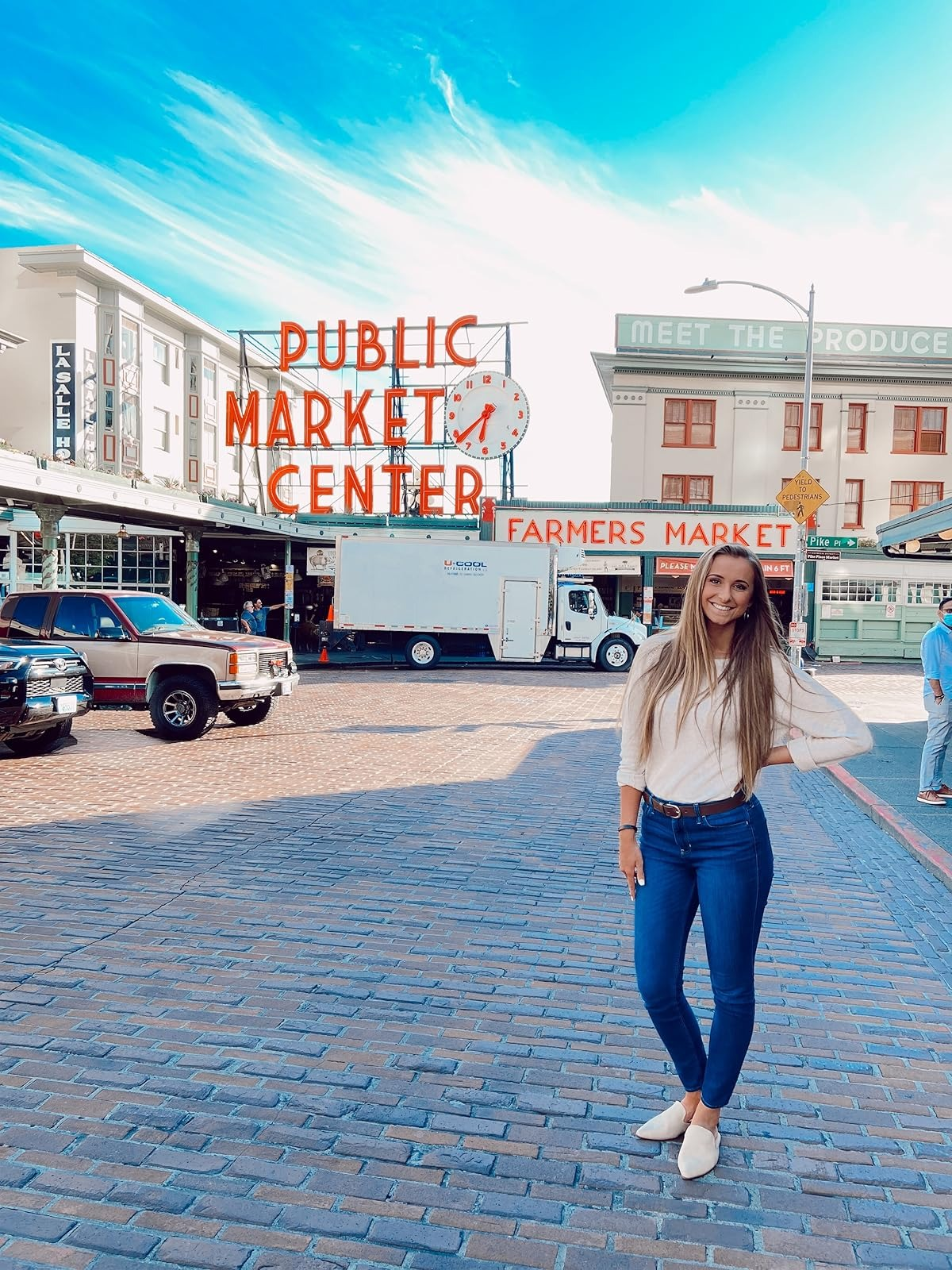 Person successful  casual outfit poses by Pike Place Market entranceway  with iconic neon sign, wearing a beige top, bluish  skinny jeans and beige suede mules