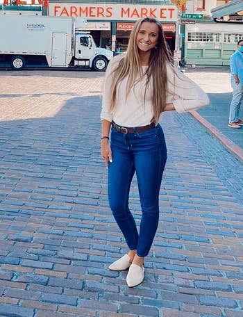 Person successful casual outfit poses by Pike Place Market entranceway with iconic neon sign, wearing a beige top, bluish skinny jeans and beige suede mules