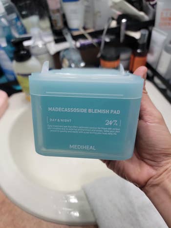 Hand holding a Mediheal Madecassoside Blemish Pad container for skin treatment with visible skincare products in background