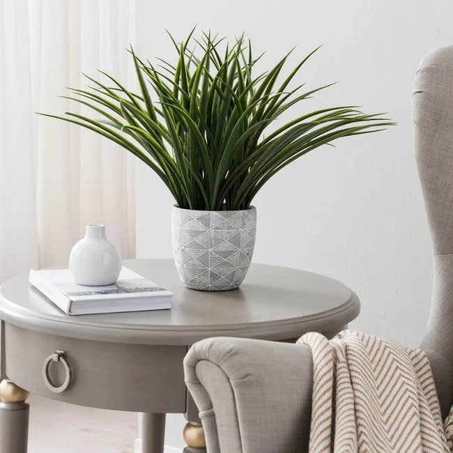 Fake green reed plant in a gray and white geometric pot on top of gray side table next to plush gray chair