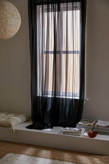 A cozy, minimalist room with a window covered by a sheer black curtain panel, a cushioned bench below, and a stack of magazines and a red glass vase on a ledge