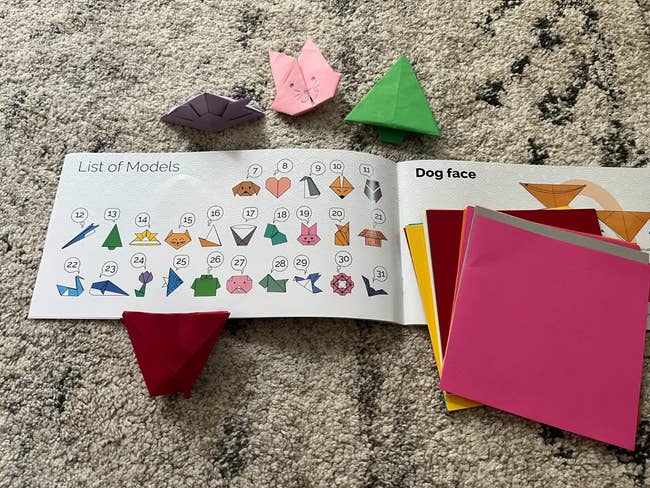 Origami book with a list of model diagrams, colorful folded shapes, and sheets of origami paper on a textured surface