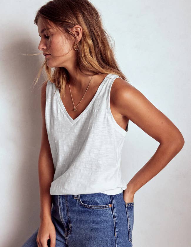 Person wearing a casual sleeveless achromatic  apical  and jeans, accessorized with a necklace and earrings, stands against a plain background