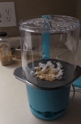 Popcorn popping inside a blue air popper in a kitchen setting, with popcorn kernels visible. Electrical outlets and kitchen counter items are in the background