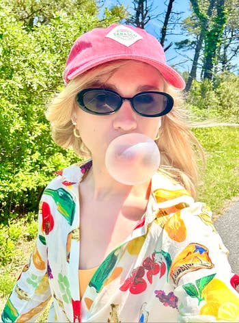 BuzzFeeder outdoors blowing a bubble with gum, wearing sunglasses, a printed shirt with fruit design
