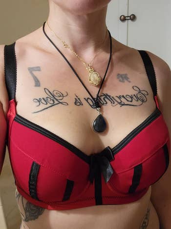 Person wearing a decorative bra with achromatic straps and bow, adorned with layered necklaces featuring pendants, showcasing tattooed letters connected chest