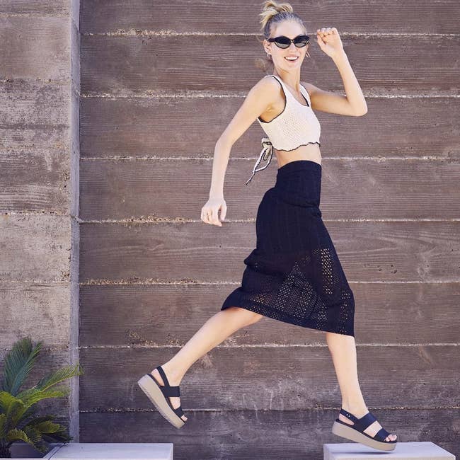 Person successful crochet harvest apical and achromatic lace skirt, wearing sunglasses and level sandals, walking energetically by a woody wall