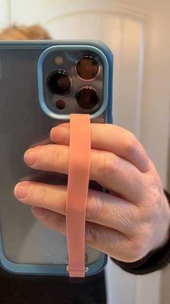 Person holding a smartphone with a blue case and pink grip strap shown in a mirror reflection