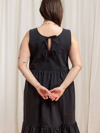 model in a casual black sleeveless dress with back tie detail and ruffled hem, viewed from behind