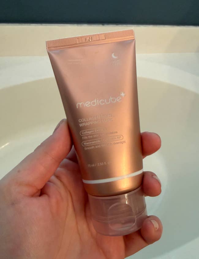 Hand holding a medicube Collagen Wrapping Mask tube, 75ml size, against a bath background