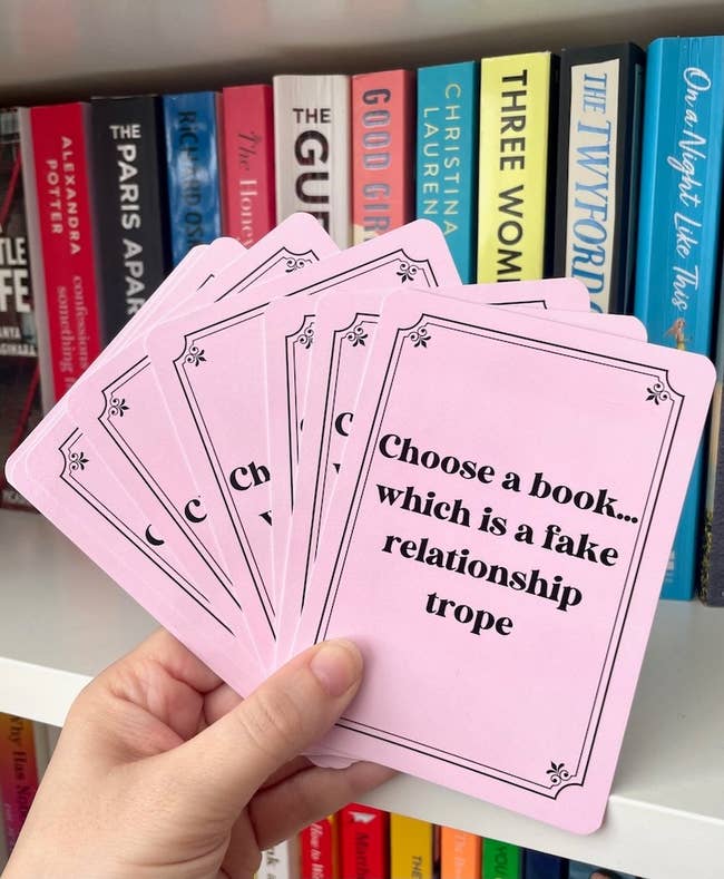 Hand holding cards with prompts for a book-related game against a bookshelf
