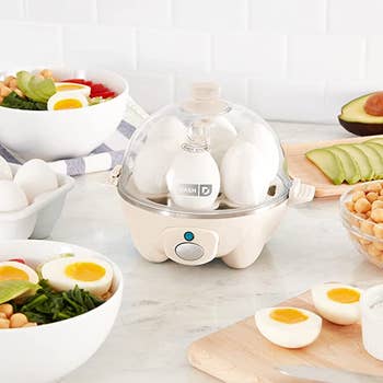 egg cooker in cream color on a counter surrounded by salads with hard boiled eggs