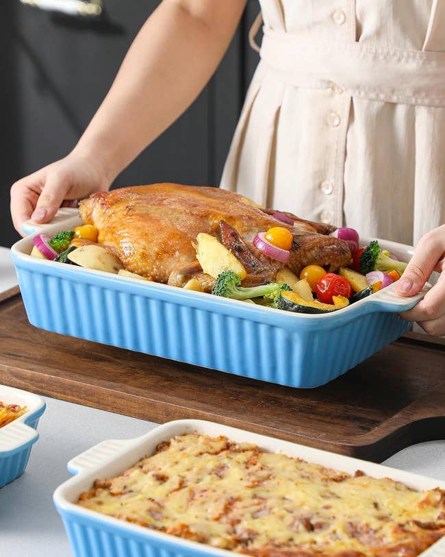 Person holding a blue dish with roasted chicken and vegetables, next to a casserole
