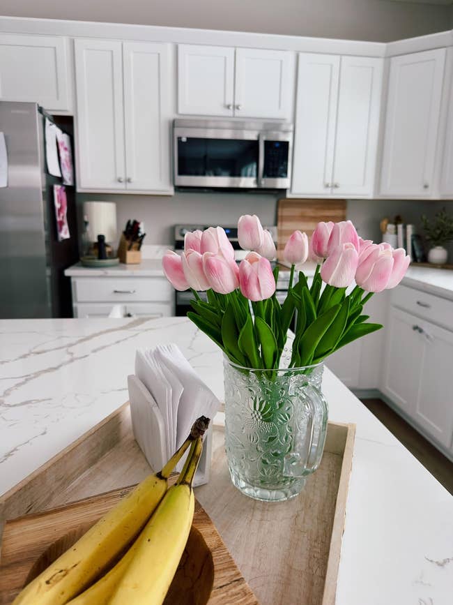 Kitchen antagonistic  with a vase of tulips, tray with bananas, and napkins. White cabinets and stainless alloy  appliances successful  the background