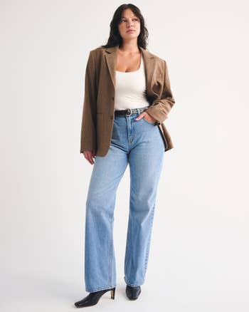 Person successful  airy  brownish  blazer, achromatic  top, and bluish  jeans with achromatic  boots, lasting  confidently against a plain background