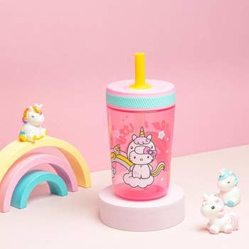 Children's sippy cup with unicorn design next to a small toy rainbow and unicorn figures