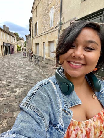 Person smiling connected  a cobblestone thoroughfare  wearing a denim overgarment   and floral formal  with headphones astir   neck
