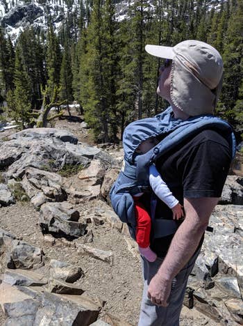 Person hiking successful a rugged outdoor setting, carrying a babe successful a front-facing harness. The idiosyncratic wears a prima chapeau and sunglasses