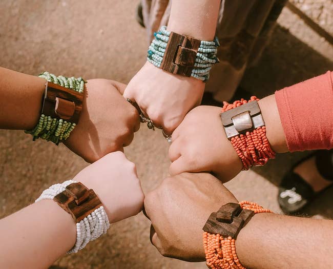 Five models wearing stacked beaded bracelets in white, orange, red, blue, and green geld together with a wooden clasp