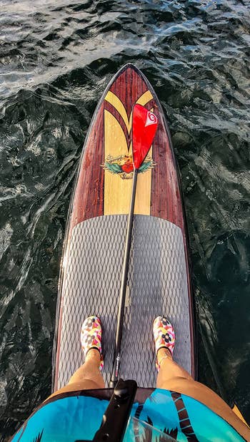 Person paddleboarding connected water, presumption from above. Colorful shoes visible. Paddle with reddish leaf placed crossed the board