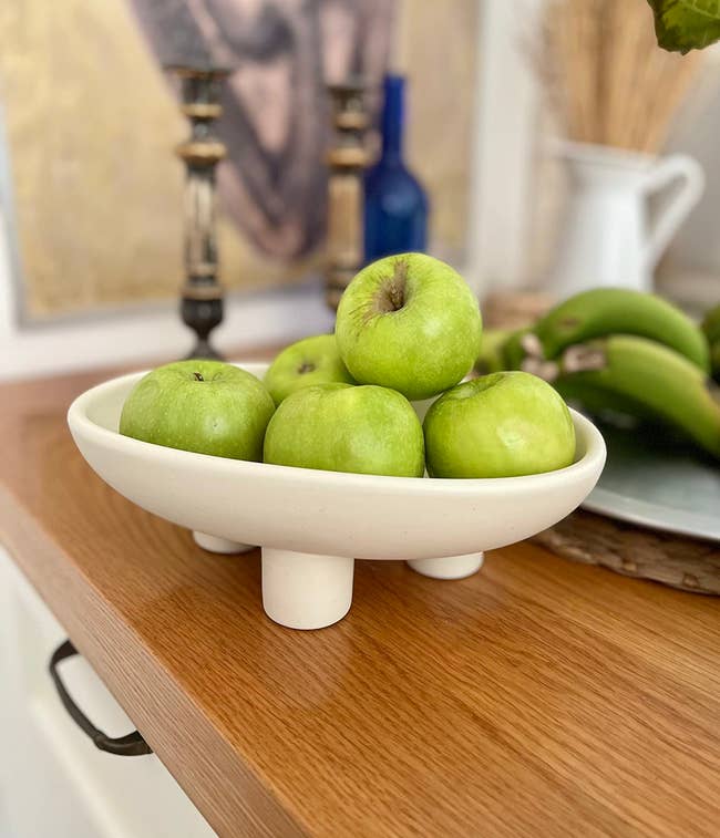 A bowl of green apples sits on a wooden counter, with a blurred background including a painting, candlesticks, a blue bottle, and a white pitcher