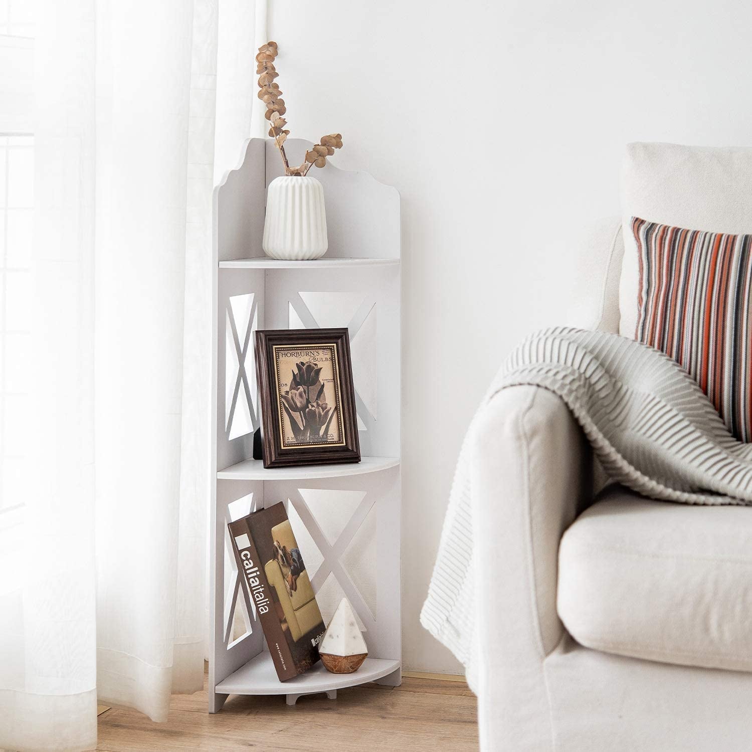 A white corner shelving unit with three shelves holding pictures, books, and a faux plant next to a couch 