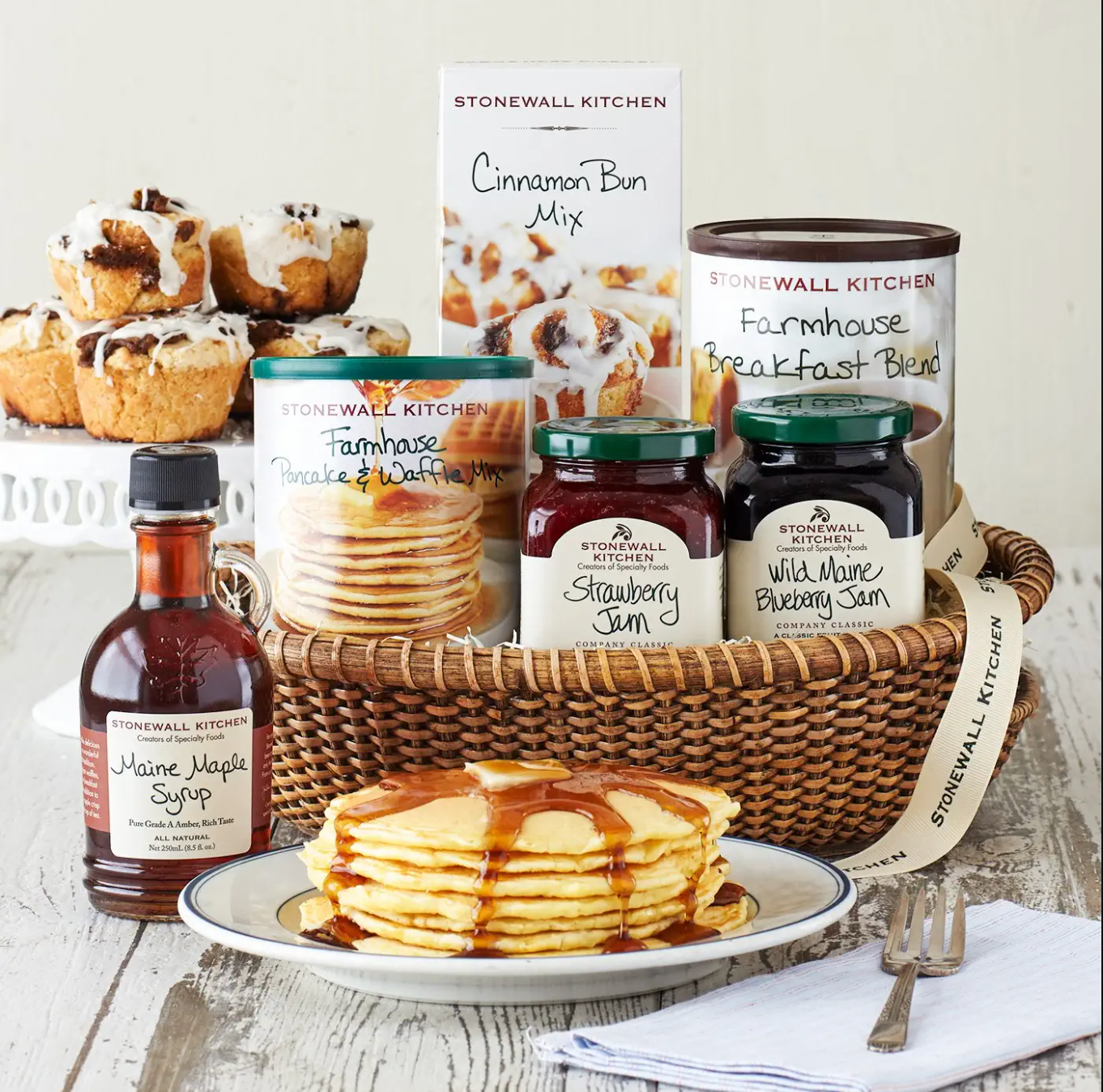 Brown wicker basket with two jars of jam, a container of pancake mix, a box of cinnamon bun mix, a container of coffee, behind a bottle of maple syrup and plate of stacked pancakes